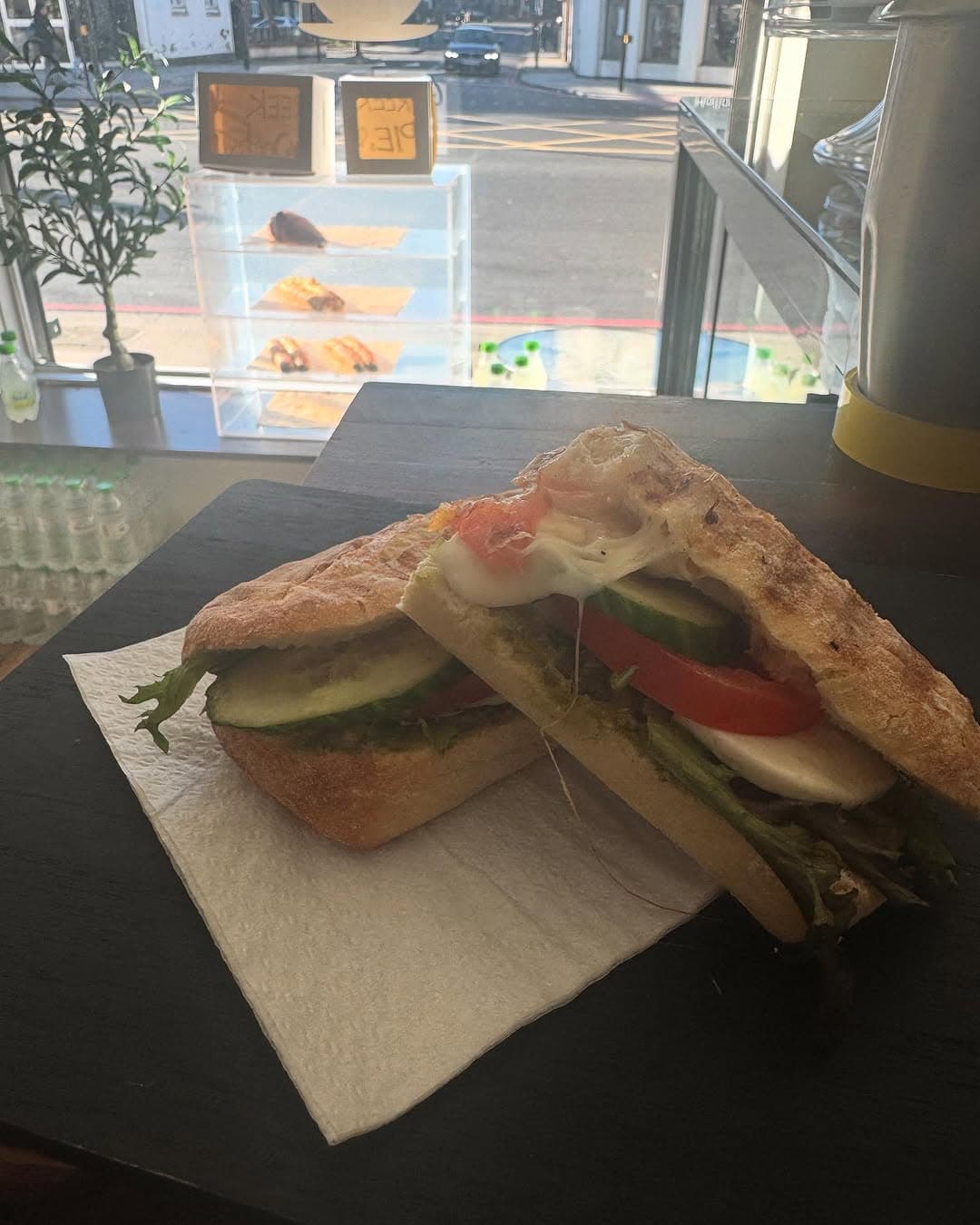 A caprese ciabatta on dark slate at the window, the chalked Greek Pies sign behind