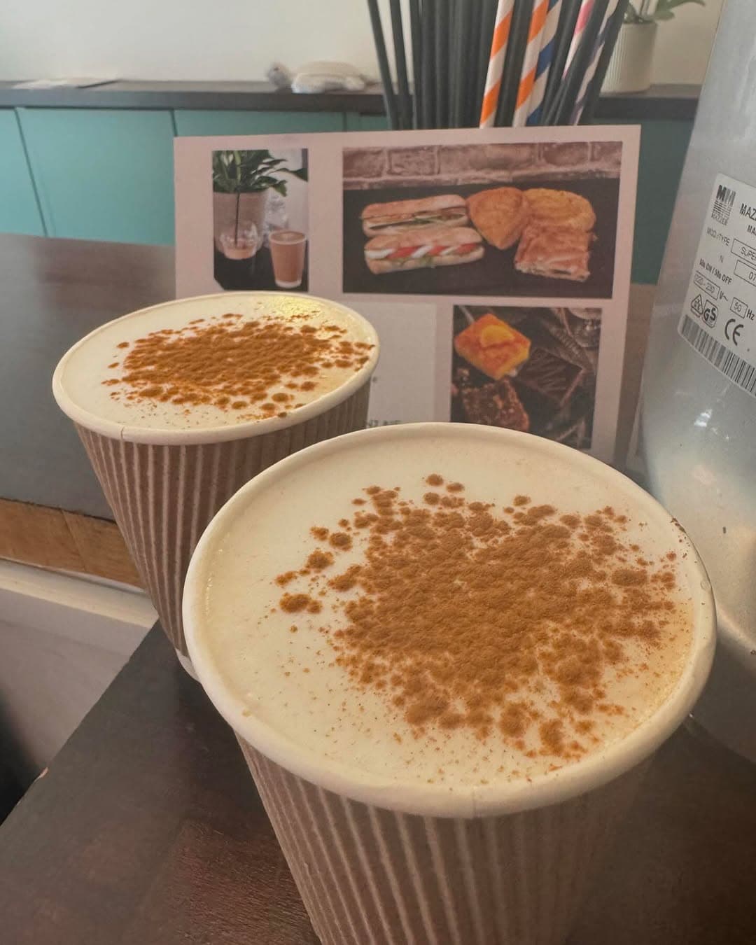Two cinnamon-dusted freddo coffees in ripple cups on a dark counter, the teal cabinet behind